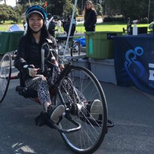 A woman sits on an adaptive recumbent bike in Golden Gate Park; a sign for BORP is in the background.