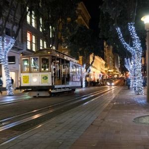 A Cable Car on a city street lined with buildings, shops and trees decorated with lights