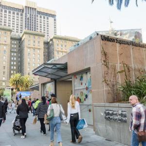 Street view of a station entrance and downtown buildings with dozens of pedestrians holding various items 