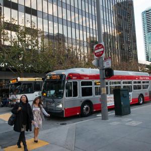 People crossing a downtown street in front of Muni and Golden Gate Transit buses