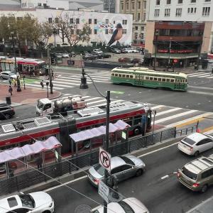 Image of a busy intersection corner possibly during commute showing multiple cars and buses, pedestrians and a trolley 