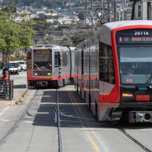 Photo of an older Breda model Muni Metro train on the street in the background, and a new LRV4 model Muni Metro train in the for