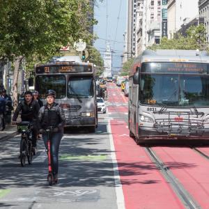 Two buses, a bicyclist, a pedestrian and person riding a scooter all on Market Street