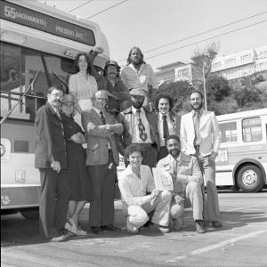 A dozen people dressed in business casual posting for a group photo in front of a bus in what seems to be a bus yard.  They are 
