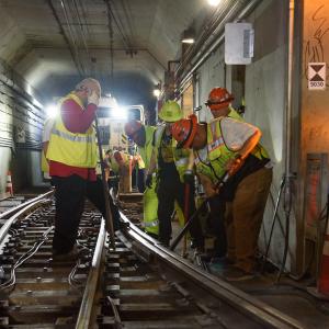 Several crew in safety gear standing over a rail track with tools angled in hand. 