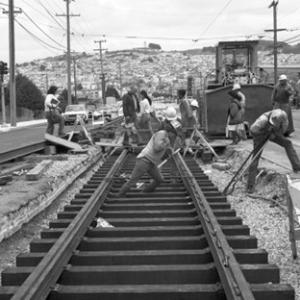Group of people working on tracks, one pushes a rail while the other lifts a tie into place
