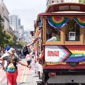 People march down the street at a parade around a historic cable car with rainbow flags.