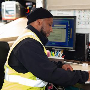 A photo of a Muni inspector writing on a clipboard at a desk with a computer and Muni Metro trains operating behind him.