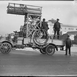 A black and white image of an old truck with a man in the driver seat, two in the in the truck bed, and one standing behind