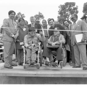 Black and white photo of people at a ribbon cutting ceremony. Two men in wheelchairs are in front to cut the ribbon.