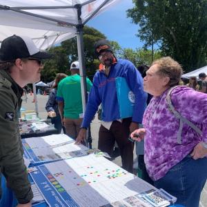 People talk underneath a tent with charts on a table and more people and tents in the background.