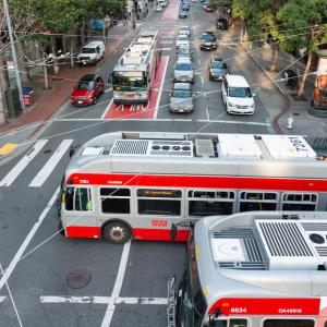 Buses and cars at an intersection with overhead wires.