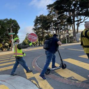 Three people in an intersection. One holding a stop sign, one on a scooter and one with a baby. 