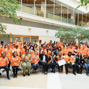 A group of SFMTA staff wearing orange and smile and wave to a camera.