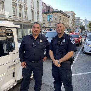 Two Parking Control Officers stand on the side of a busy street with cars and buildings in the background.