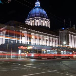 A night time photo of San Francisco City Hall with a Muni bus driving by.