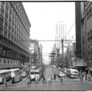 A black and white photo of a busy street with buses, cars and people crossing at a cross-walk.