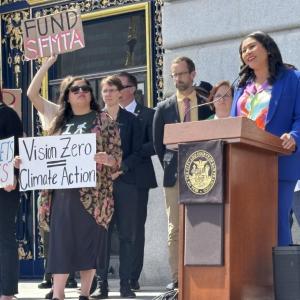 Mayor Breed speaks at a podium in front of city hall. People stand near her with signs that say Fund SFMTA and Vision Zero.