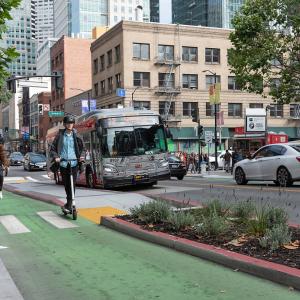 A person rides a scooter in the protected 2nd Street bike lane alongside a cyclist. A Muni bus is visible in the background.