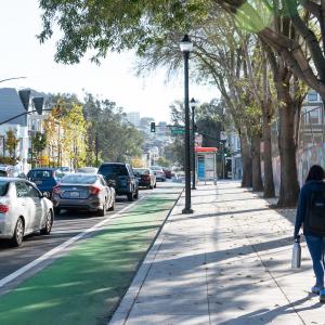 Woman walks on a sidewalk toward a bus shelter. On her left is a green bike lane beside lanes of cars.
