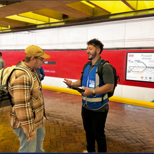 Survey taker wearing a blue vest and badge speaks with a Muni rider on the Powell Station platform for the onboard ridership sur