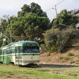 Historic streetcar painted cream and green moves through Dolores Park with a bridge in the background and grass near tracks.