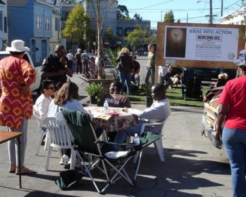 People gather at tables at a street meeting. Poster reads "Ideas into action".