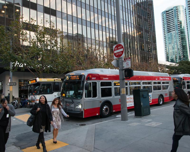 People crossing a downtown street in front of Muni and Golden Gate Transit buses