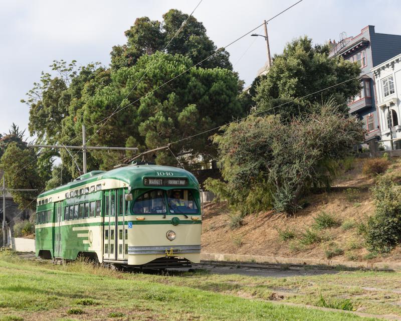 Historic streetcar painted cream and green moves through Dolores Park with a bridge in the background and grass near tracks.
